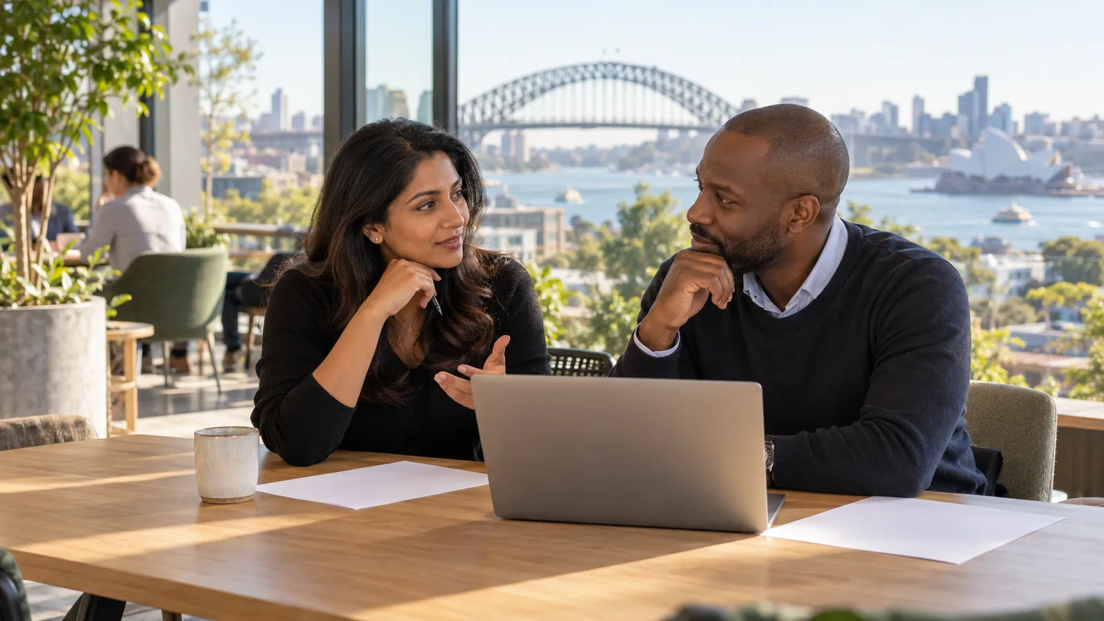 Two professionals discuss plans beside a laptop in a sunlit workspace overlooking Sydney Harbour Bridge.